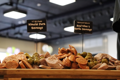 A side view of a display with two kinds of sliced sausages intermingled with piles of green olives. Two signs stick out at the top of the display. One reads ""VT 99 Meats Kimchi Pork Sausage." The other reads "Vermont Salume Red Wine and Garlic Sausage."