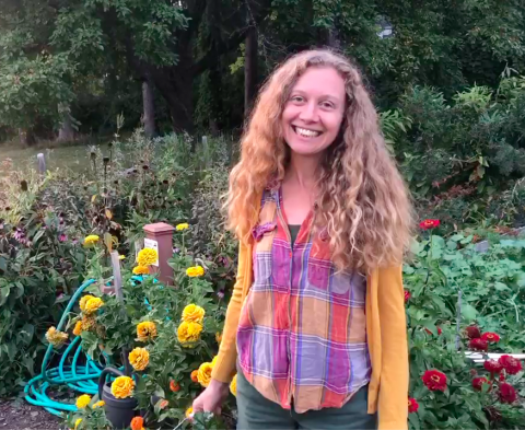 A smiling woman with curly dark blonde hair stands in a garden. She is wearing a button-down shirt with large orange, blue and purple checks and yellow sleeves.