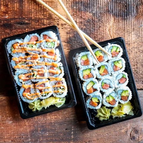 Two black containers sit on a rustic wooden table. Both contain 12 slices of sushi rolls, each with pink salmon and green avocado inside, surrounded by white rice. At the bottom end of each container there is a dollop of green wasabi and a nugget of pickled ginger. Two bamboo chopsticks are resting on the top of the second container.