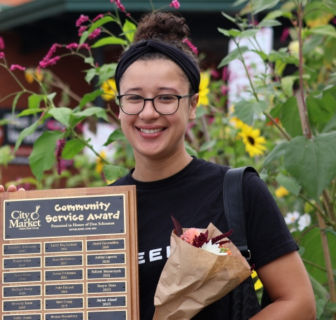 A photo of a person from the waist up. She is holding a plaque that reads "Community Service Award" at the top and a bouquet of flowers. She is wearing a black tee shirt, a black headband, and black-rimmed glasses. She is wearing her hair in a bun at the top of her head.