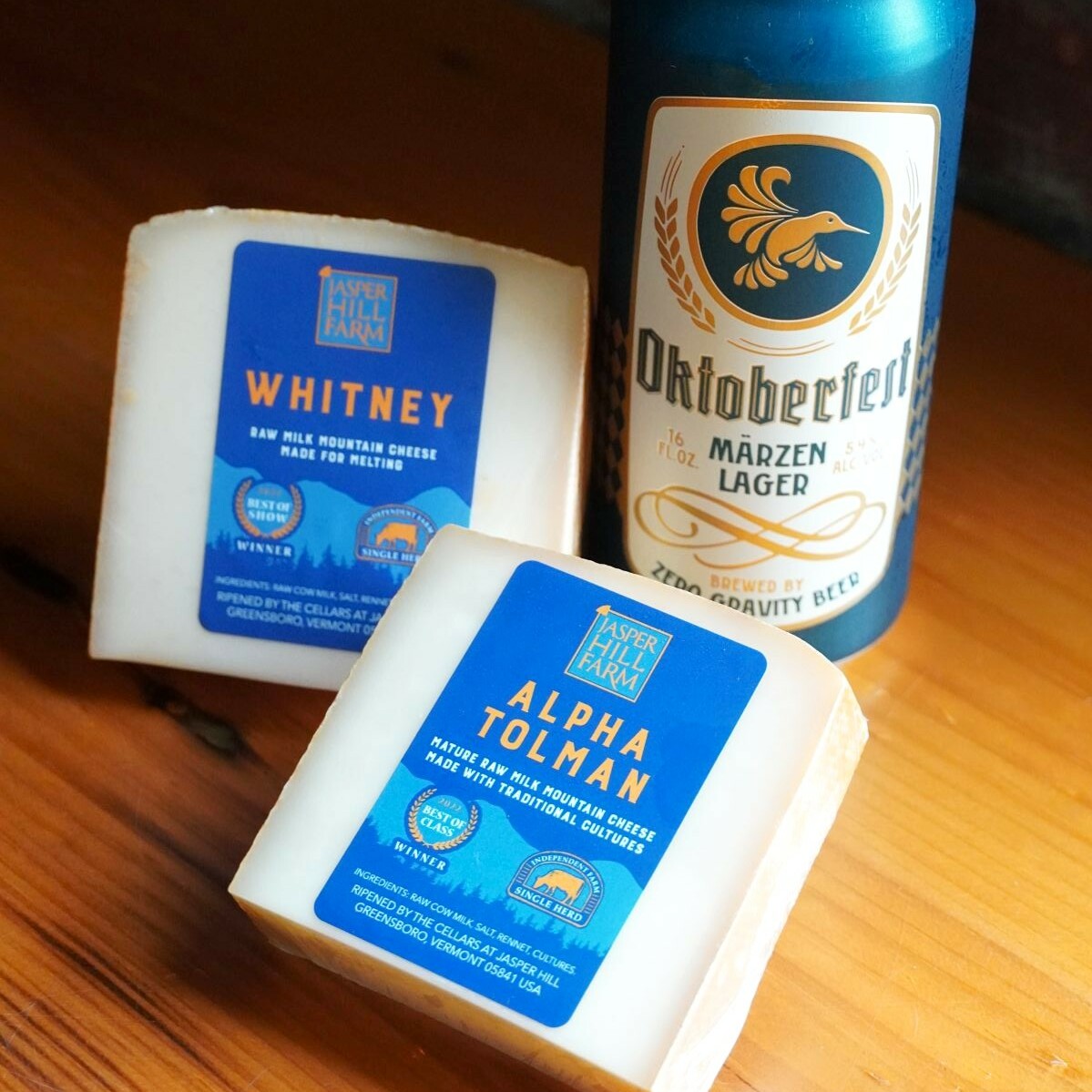 Two cheese blocks labeled "Whitney" and "Alpha Tolman" next to a can of Zero Gravity "Oktoberfest" beer.