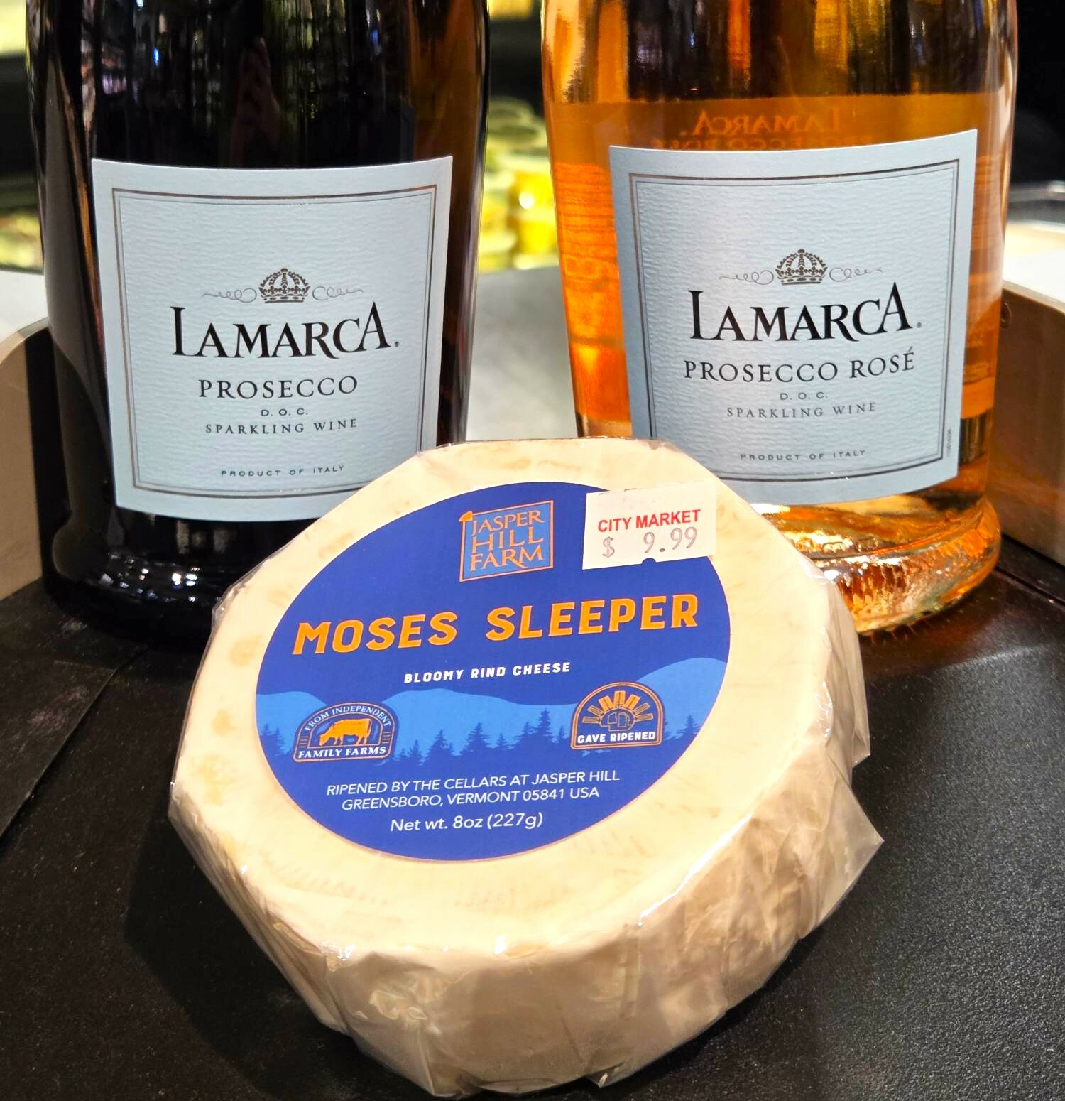 Two bottles of La Marca Prosecco and a round of Moses Sleeper cheese on display.