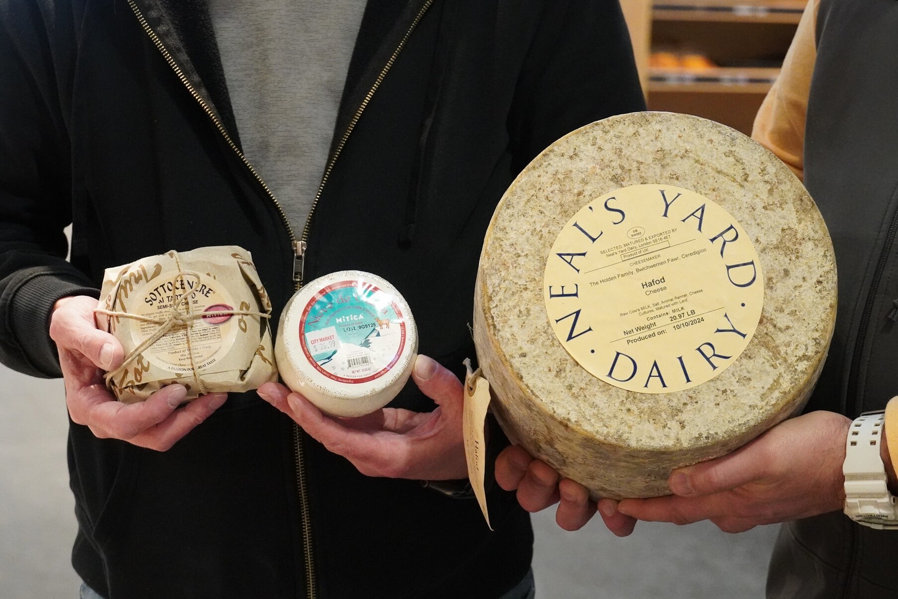 Three types of cheese being held: one wrapped, one labeled "Mitica," and a large round from Neal's Yard Dairy.