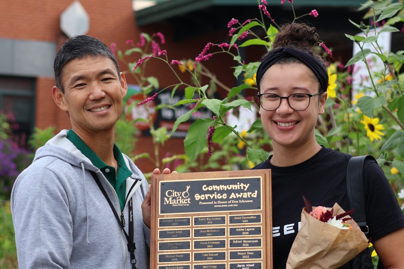 Two smiling individuals pose with a community service award in a garden setting.