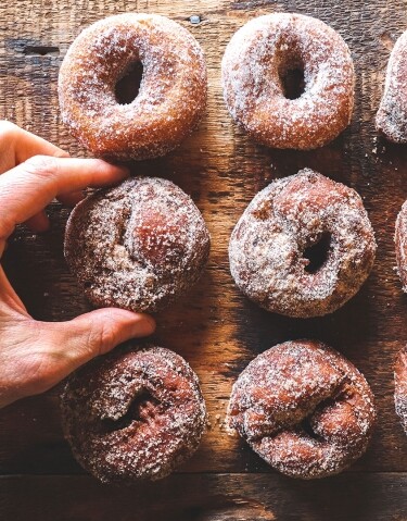 A hand selecting a sugared donut from a wooden surface with several donuts arranged.