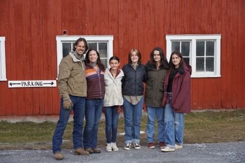 A man and five women stand together in front of a red barn.