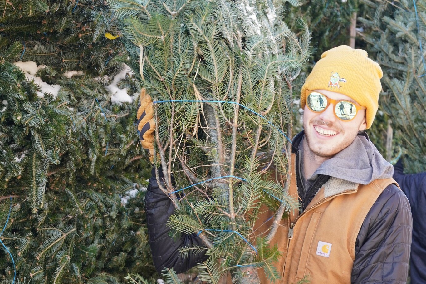 Person in sunglasses and beanie smiles while holding a Christmas tree.