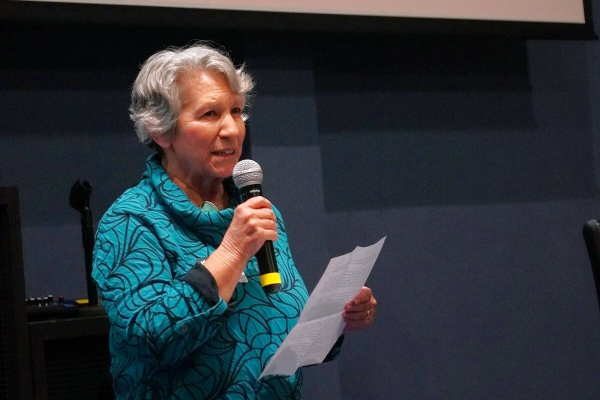 a woman stands with a microphone and paper in hand talking to a crowd