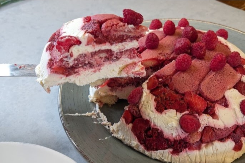A slice of layered berry and cream cake is being lifted from the full cake.