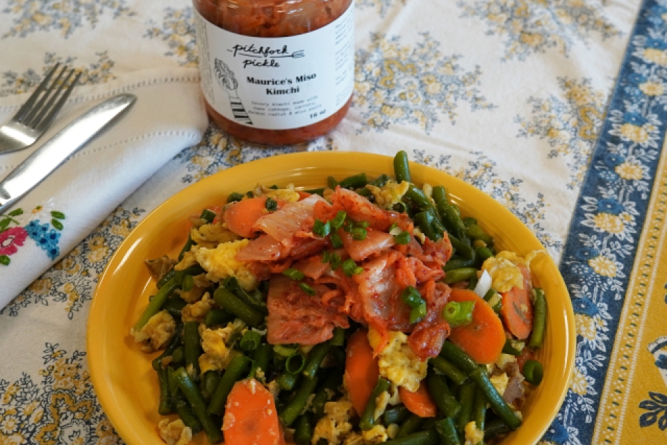 On top of a floral tablecloth sits a yellow plate filled to the brim with carrots, eggs, scapes, scallion, kimchi and other veggies. Behind the plate is an open jar of Pitchfork Pickle's kimchi. To the left is a folded napkip with a fork and knife. 