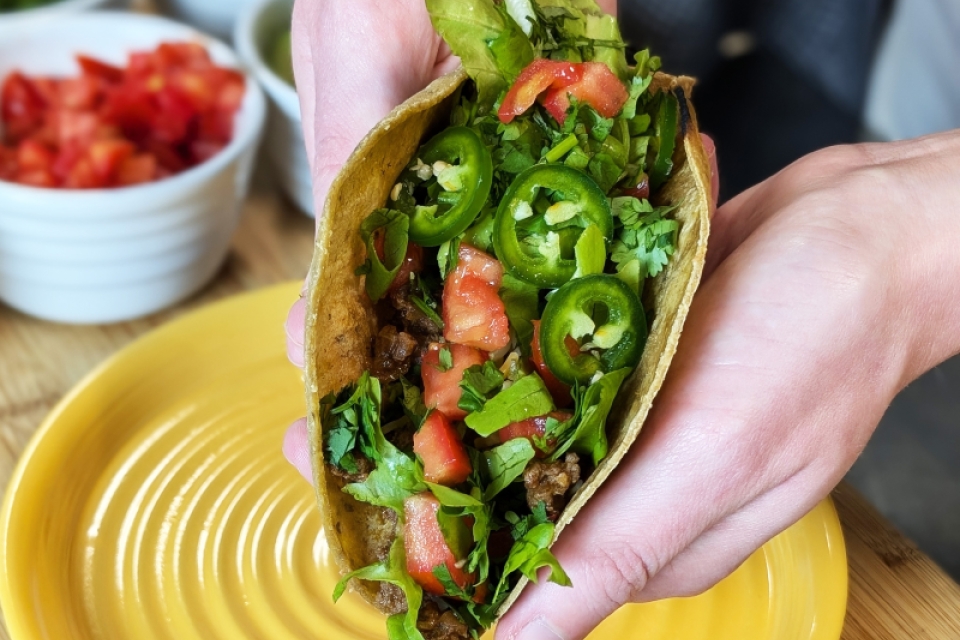 Two hands cradle a soft-shell taco. It is topped with bright green jalapenos, darker green cilantro, and bright red tomato.