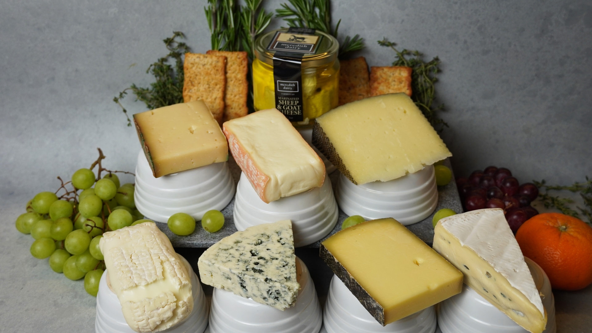 Seven cheeses are arranged on short white pedestals. They are surrounded by laurels made of fruit, herbs and crackers.