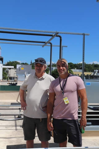 Two people stand on a roof. The first is wearing a white polo shirt and shorts. The other is wearing a pink tee shirt, a lanyard with a rectangular card on it, and shorts. Behind them, the roof is studded with ventilation equipment. The sky behind the people and equipment is a rich blue.