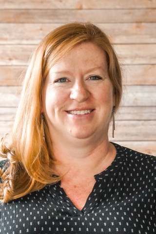 A photo of a smiling person from the shoulders up. The person is wearing a black shirt dotted with tiny white chevrons. She has shoulder-length red hair tucked over her right shoulder and bangs.