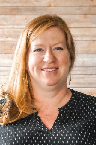 A photo of a smiling person from the shoulders up in front of a wood-panel backdrop. The person is wearing a black shirt dotted with tiny white chevrons. She has red hair pulled over her left shoulder.