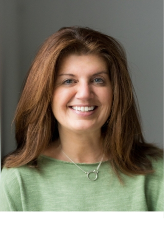 A photo of a person from the chest up. The person is wearing a light green boatneck shirt and a silver necklace. They have dark auburn shoulder-length hair. They are smiling.