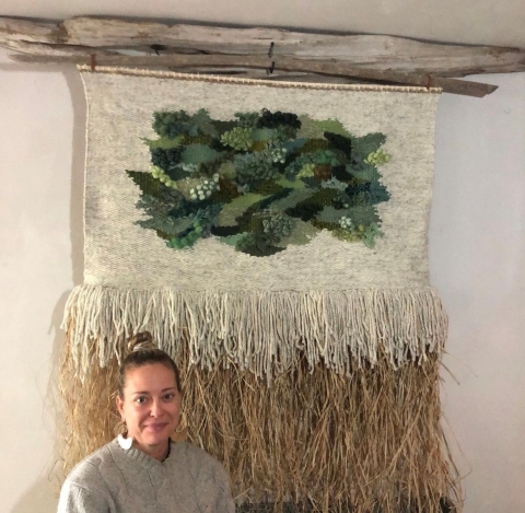A beige tapestry with several shades of green woven into the center. It is attached to a tree branch. The bottom half of the tapestry is brown and straw like. The artist sits below it, with her hair up, silver earrings, and a grey sweater.
