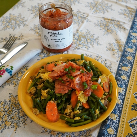 On top of a floral tablecloth sits a yellow plate filled to the brim with carrots, eggs, scapes, scallion, kimchi and other veggies. Behind the plate is an open jar of Pitchfork Pickle's kimchi. To the left is a folded napkip with a fork and knife. 