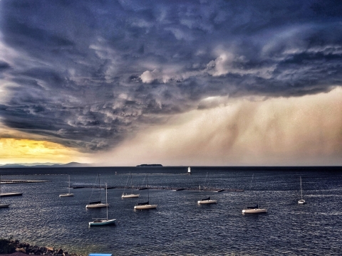 A photograph of a storm rolling in from the west over Lake Champlain. Sailboats dot the dark blue water and clouds roil the sky. In the far left of the photograph, a corner of a bright yellow sunset peeks through.