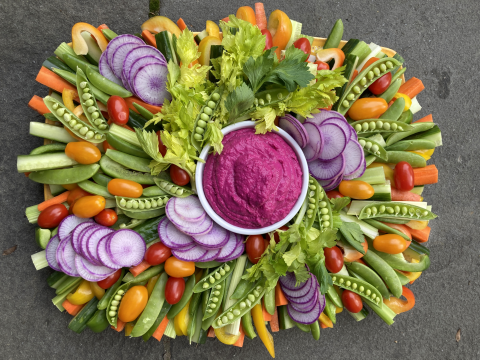 A bountiful collection of sliced vegetables, including snap peas, tomatoes, carrots, daikon, yellow bell peppers, parsley, and celery are arranged around a bowl of bright magenta beet hummus.