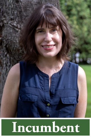 A photo of a woman from the waist up. She is standing in front of a wide, dark brown tree. She has dark brown hair and bangs, and she is wearing a dark blue sleeveless button-down blouse. She is smiling.