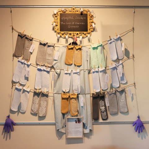 15 pairs of socks hang from wire in 3 rows of 5, with a handwritten sign above reading "Hospital Socks." Each pair has a price tag for a hospital bill.