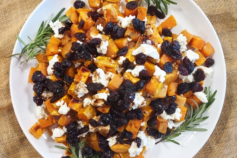 A white plate centered on a burlap table covering. The plate holds a pile of diced roasted sweet potato garnished with dark red cranberries, bright white chevre, and fresh green sprigs of rosemary.