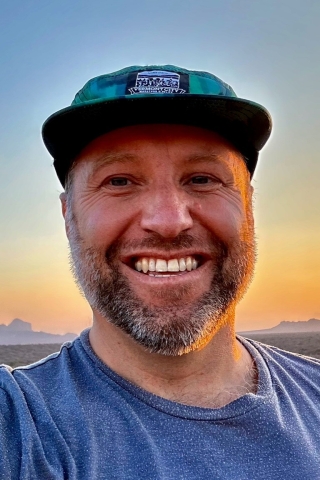 A photo of a person from the shoulders up. He is wearing a blue tee shirt and a teal trucker hat. He is standing in front of a sunset over some mountains.