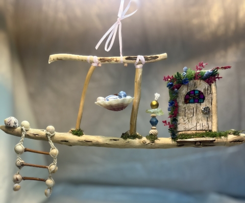 A fairy house. There is a horizontal stick, with a small ladder hanging down from the left end of it. On top of the stick are two sticks in a rectangular arch with a bow on the top. To the right are a few stacked beads, and to the right of that is a little door. The background is blueish gray.