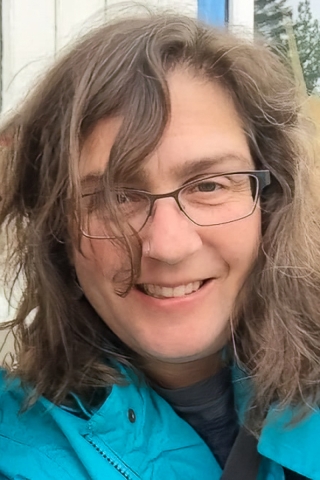 A photo of a woman from the neck up. She is wearing a bright cerulean jacket and black-framed glasses. She has brown wavy hair and she is smiling.