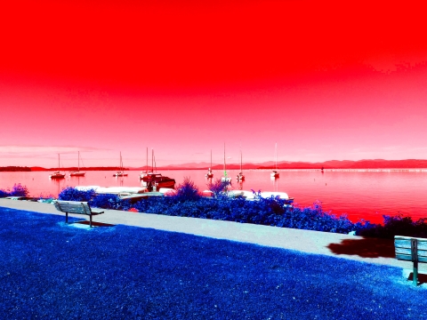 A saturated image of the waterfront. In the foreground is blue grass and light blue benches along the path. In the background, the sky, lake, and mountains are red. There are a few white boats on the lake.