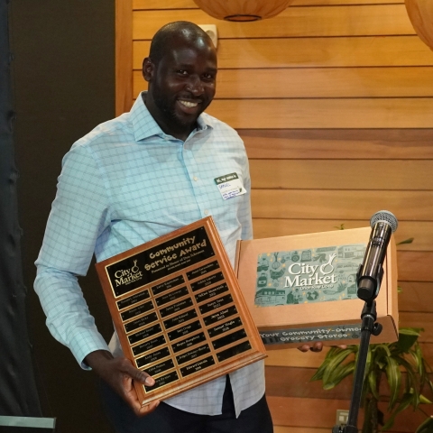 A photograph of a man holding a plaque and a gift box. He is smiling and wearing a light blue button down shirt.